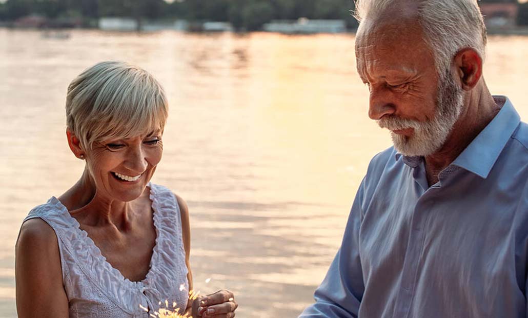 A senior couple standing waterside, holding a lit sparkler and smiling.